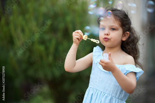 Young girl in a blue dress blowing soap bubbles in a park, enjoying playful moments of childhood innocence and fun