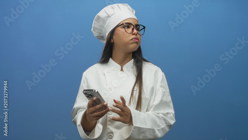 Cuadro en lienzo Young chef woman wearing white jacket and glasses holding smartphone with visible hand, glancing upward toward left in studio; curiosity