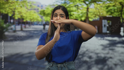 Schilderij op canvas Young hispanic woman forms timeout gesture with hands and forearms visible while standing in a tree lined urban street plaza wearing blue shirt and jeans; assertive pause
