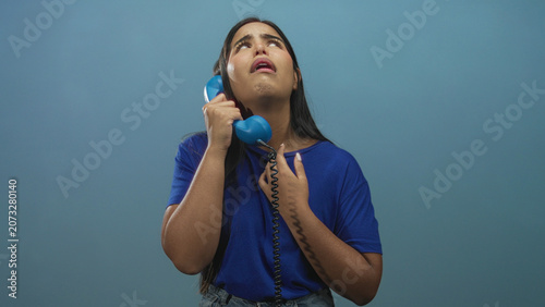 Foto Young hispanic woman holding blue corded telephone to her ear with hand raised in pleading gesture in studio; despair longing