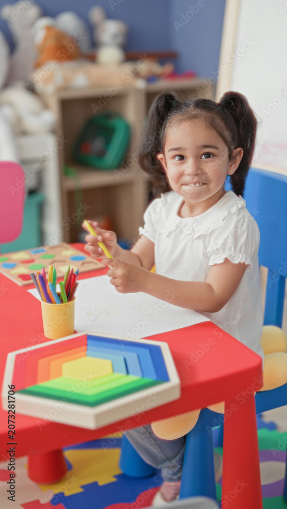 Obraz premium Girl drawing with yellow pencil at a red children's table, colored pencils cup and rainbow wooden stacking toy visible in preschool building; concentration creativity.