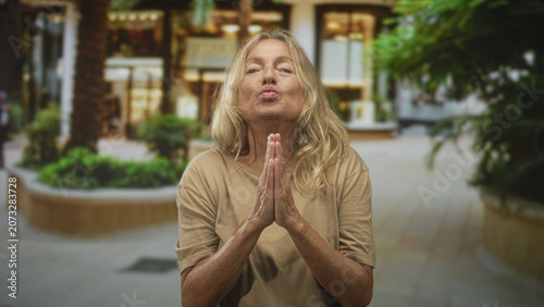 Fotografie Senior blonde woman with hands pressed together in prayer, pursed lips and closed eyes standing near a building entrance lined with plants; gratitude serenity reflection