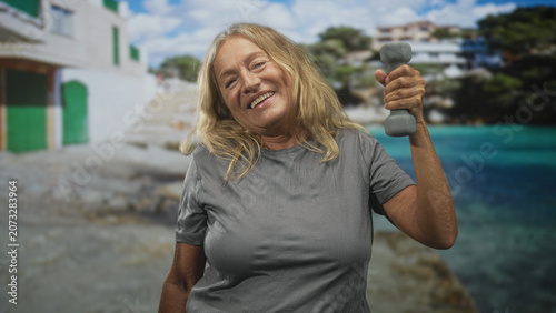 Fotografie Woman holding dumbbell aloft with right arm and hand in front of a building by the seaside; resilience wellness