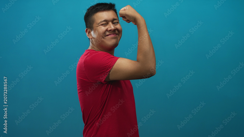 Fototapeta premium Hispanic man flexes bicep and smiles wearing red t shirt, side pose in studio with teal backdrop; confidence strength.