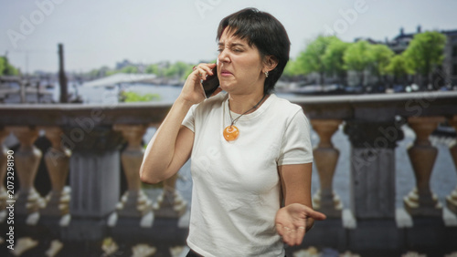Woman on street holding phone to ear, grimacing and shouting with palm up gesture while wearing a white t shirt and orange pendant; frustration.