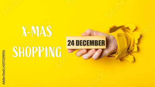Hand holding a wooden block with the date 24 December, surrounded by yellow background and torn paper, conveying festive spirit of Christmas shopping season