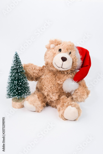 Plush teddy bear wearing a red Santa hat sits beside a small Christmas tree, creating a festive and cheerful holiday atmosphere for Christmas and New Year celebrations