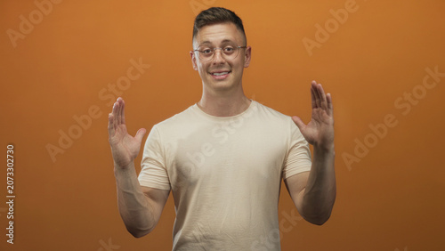 Bild auf Leinwand Young man in glasses holds hands apart palms facing outward showing space in orange studio; amused confidence