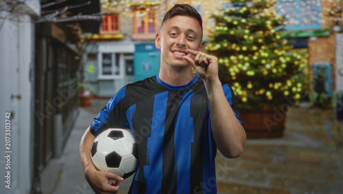 Man holding soccer ball under arm on street in front of decorated christmas tree and storefronts, wearing blue striped jersey and smiling while glancing sideways; holiday pride.