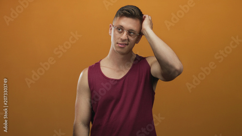 Foto Man in burgundy tank top with glasses scratches back of head showing bicep and exposed skin in studio; awkwardness uncertainty