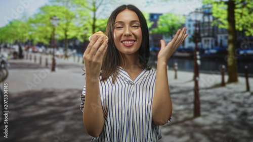 Fotografie Young hispanic woman tasting a muffin on a sunny city street, hand near mouth and smiling; joy indulgence