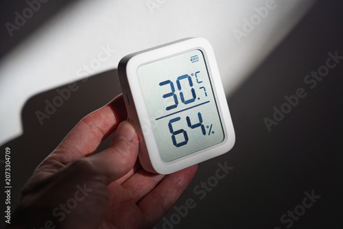Hand holding digital thermometer showing 30 degrees Celsius indoors during a summer heat wave