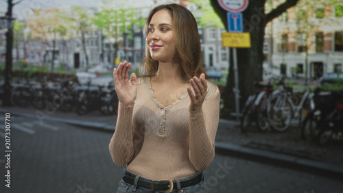 Photography Woman clapping hands on a city street in a casual sweater and jeans, smiling toward camera near bicycles and canal, belt buckle visible; joy