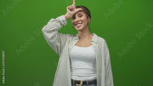 Foto Woman makes l sign with hand on forehead, smiling while wearing white tank top and striped overshirt in a green screen studio; playful teasing