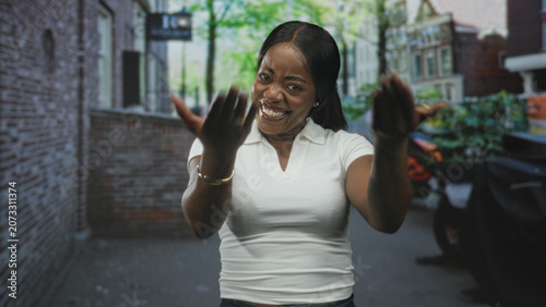 Εκτύπωση καμβά Woman smiling broadly with hands on hips on a city street in front of brick building, wearing white shirt and bracelet; joyful confidence