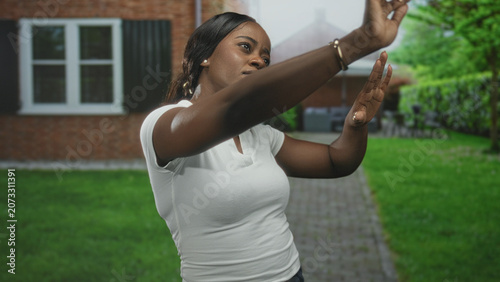 Quadro su tela Woman with raised hands and visible forearms shielding or framing a view, standing in front of a brick building and lawn; determination focus