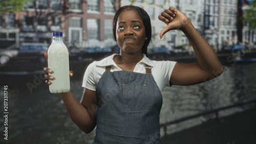 Cuadro en lienzo Woman in apron holds milk bottle with one hand and shows thumb down gesture in studio; disapproval concern