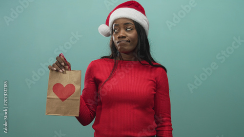 Fényképezés Woman in santa hat and red sweater holds brown paper gift bag with red heart in one hand, shrugs and makes unimpressed face in studio; holiday doubt