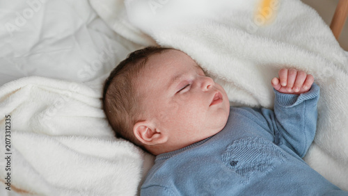 Fotografía Newborn boy sleeping in a blue sweater with raised hand on a soft white blanket in a nursery cradle, eyes closed and relaxed; quiet serenity
