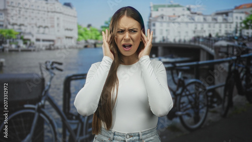 Фотография Woman covering ears with hands beside parked bicycle on urban street near canal bridge wearing white shirt and jeans; annoyance coping