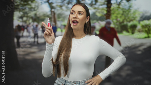 Fotografía Woman holding vaporizer near mouth, index finger raised, standing on a park path with trees and blurred pedestrians; confidence casual leisure