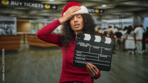 Fotografie Woman in santa hat holding a clapboard with eyes closed and hand pressed to forehead in an airport ticketing hall with crowd and buy tickets sign visible; holiday stress