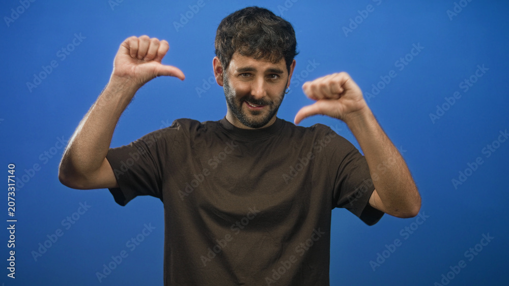 Fototapeta premium Man points thumbs to chest in studio against blue backdrop, bare thumbs and forearms visible, beard and slight smile while facing camera; confidence.