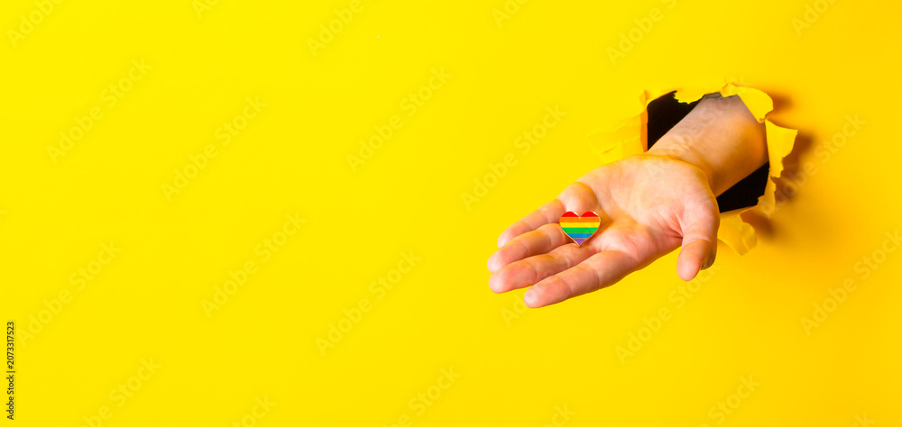 Fototapeta premium Hand Holding Rainbow Heart Through Yellow Paper Background