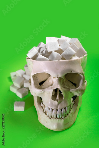 Toma frontal a nivel de los ojos de un craneo humano artificial desbordando cubos de azucar blanca sobre un fondo verde solido con terrones apilados a un lado.