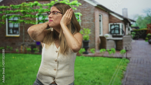 фотографія Woman covers ears with hands in front of building patio outside a house, wearing glasses and vest, displaying a startled reaction and tense expression; noise anxiety
