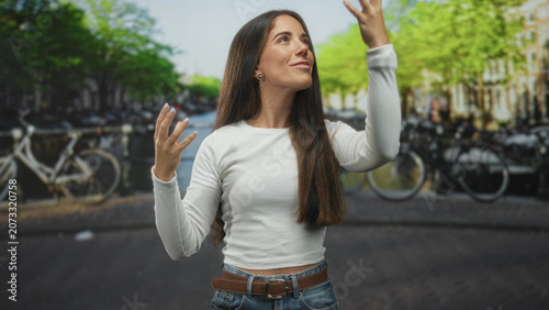 Fényképezés Young woman with hand to nose and expressive gesture on a street by a canal lined with bicycles; playful curiosity