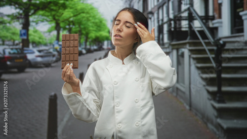 Bild auf Leinwand Woman chef holding chocolate bar, touching forehead with hand while wearing white coat on a street lined with townhouses; fatigue reflection