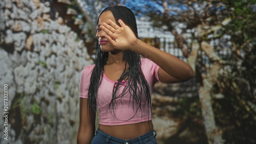 Bild auf Leinwand Woman covering eyes with hand on street by stone wall wearing pink crop top and jeans; privacy shyness