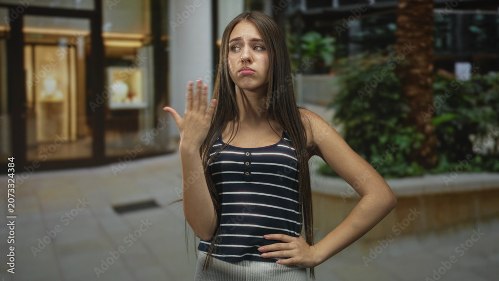 Fototapeta premium Teenage girl with long hair holding hand near mouth in a dismissive pout gesture inside a building, palm raised and one hand on hip; indifference attitude.