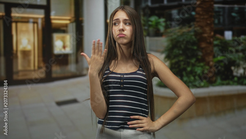 Foto Teenage girl with long hair holding hand near mouth in a dismissive pout gesture inside a building, palm raised and one hand on hip; indifference attitude