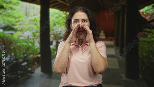 Photos Woman cupping hands to mouth calling in building veranda amid garden and wooden columns, wearing pink polo and sitting on a seat; friendly invitation