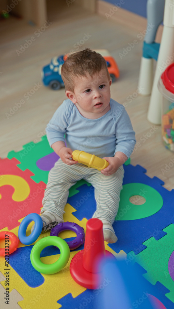 Fototapeta premium Boy holding yellow ring toy sitting on colorful foam mat in a building playroom; curiosity exploration innocence.