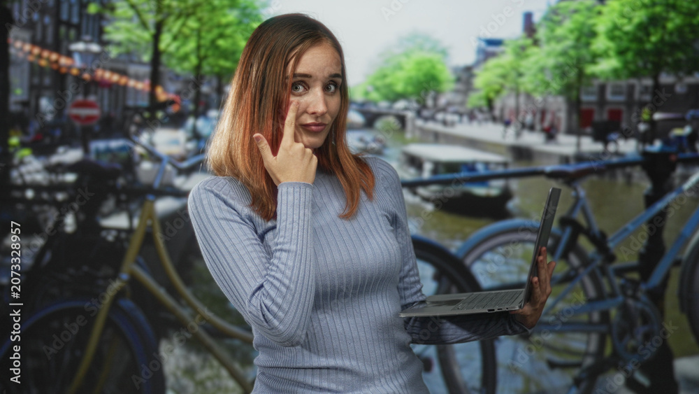 Fototapeta premium Woman with red hair holding a laptop and touching her nose with one finger while standing on a bicycle lined canal street with blurred canal boats; thoughtful remote work.