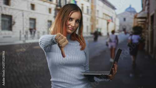 Tableau sur toile Woman holding laptop with thumb down gesture on a cobblestone street beside a historic building, redhead in a light sweater; disapproval critique feedback