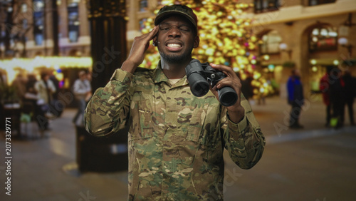 фотографія Soldier man holding binoculars, hand to temple inside a building decorated with christmas tree and lights, crowded with people; vigilance duty