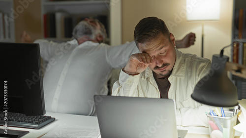 Konstfotografi Two men working late at desks in a building, one rubbing his temples and the other with hands clasped over a laptop; overnight fatigue