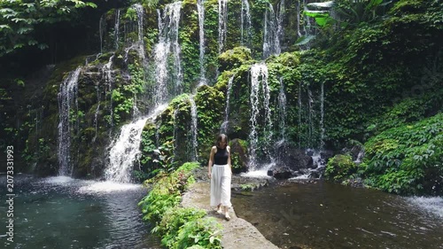 Woman walk on a path to tropical waterfall surrounded by rainforest greenery