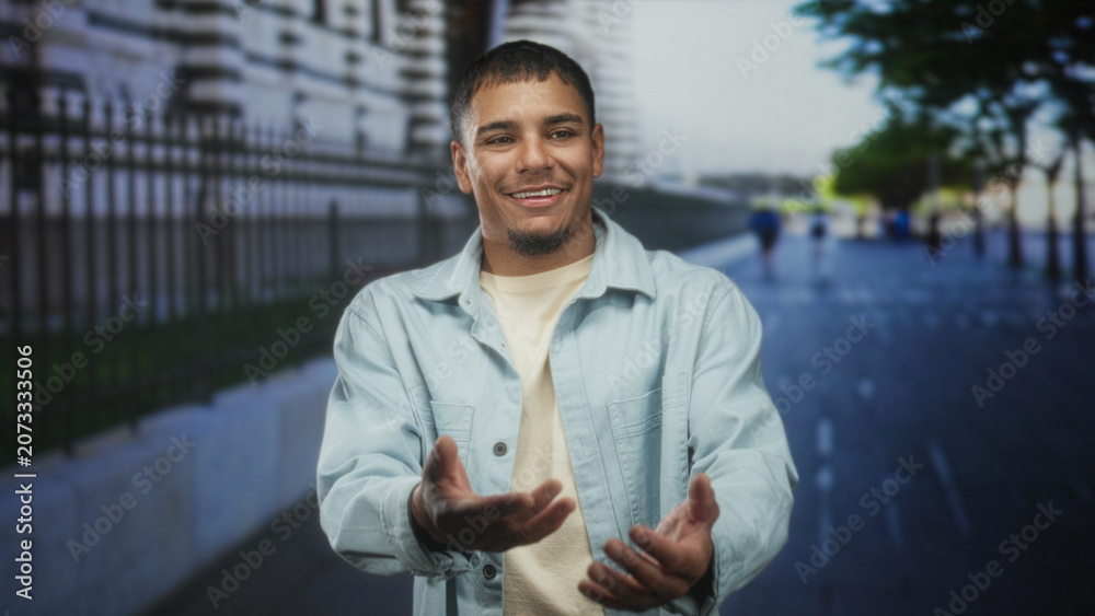 Obraz premium Man smiling, hands open and gesturing while standing on a street beside a stone wall and iron fence in front of a building; optimism approachability.