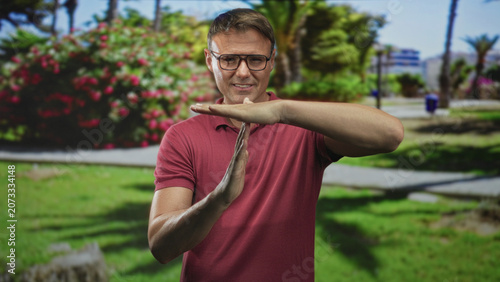 Foto Man in glasses making time out hand gesture with exposed forearms while standing on a sunny street lined with palm trees and flower bushes; caution restraint