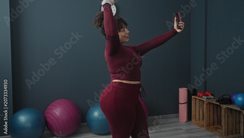 Schilderij op canvas Woman holding red smartphone aloft taking a selfie in a studio with yoga balls and dumbbells visible; confidence wellness
