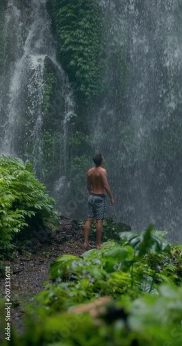 Man with raised arms under powerful waterfall, feeling energy in tropical jungle nature