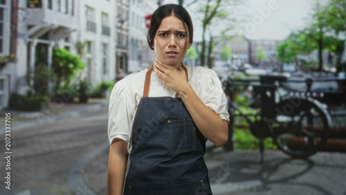 Foto Woman in denim apron holding neck with hand on street by bicycle and canal and building, pained expression; anxiety discomfort