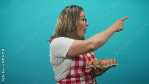 Obraz na plátně Woman in a studio wearing red check apron holding a carton of eggs and points finger to the right with a stern open mouth expression; firm disapproval