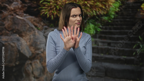 Foto Woman holding hands up palms out on stone steps wearing a blue ribbed sweater with tense frown in forest setting; disgust refusal avoidance