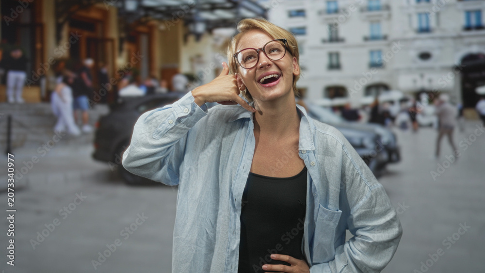 Fototapeta premium Woman making call gesture with hand to ear, wearing glasses and light blue overshirt on a busy street with cars and building facades; playful confidence.